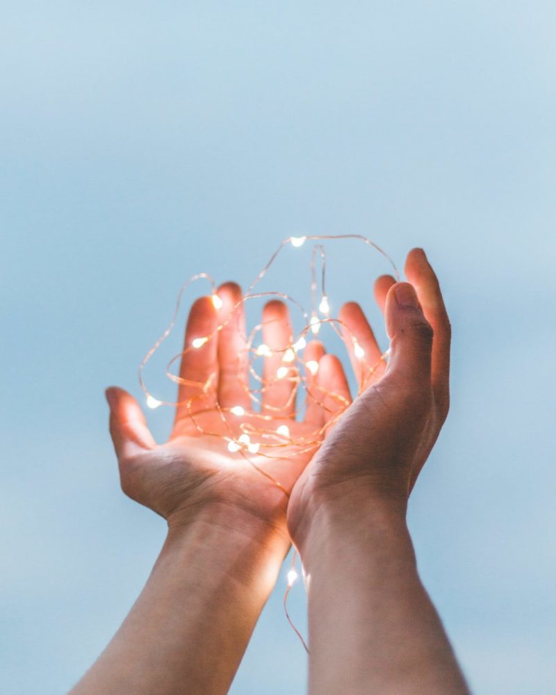 Hands holding up string lights with the sky in the background.