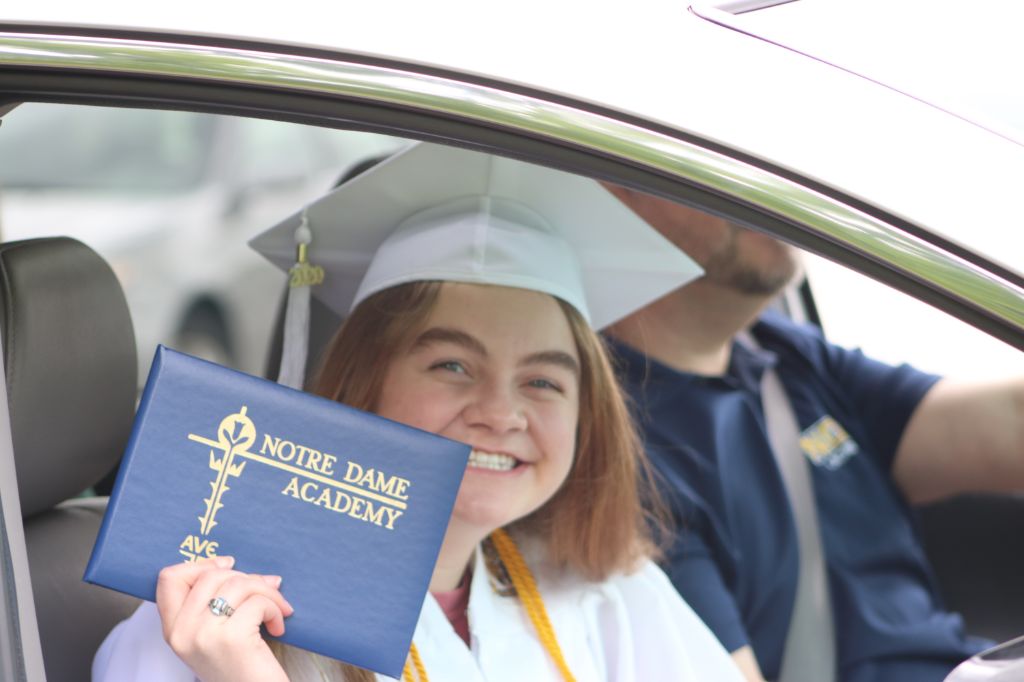 A student sitting in their car holding their diploma and smiling at the camera at the Notre Dame Academy catholic all-girls school in Covington, Northern Kentucky.